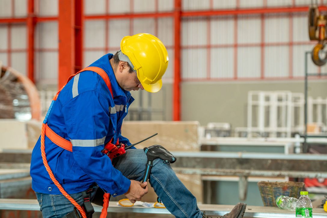 Ein Arbeiter in blauer Jacke und gelbem Schutzhelm kniet auf einer Metallkonstruktion und benutzt Werkzeuge in einem hell erleuchteten Industrielager.