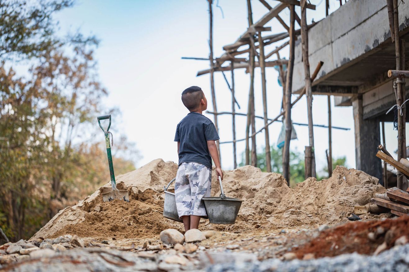 Ein kleiner Junge steht auf einer Baustelle, hält einen Eimer in der Hand und blickt auf ein teilweise fertiggestelltes Gebäude, das von Schutt und Sand umgeben ist.
