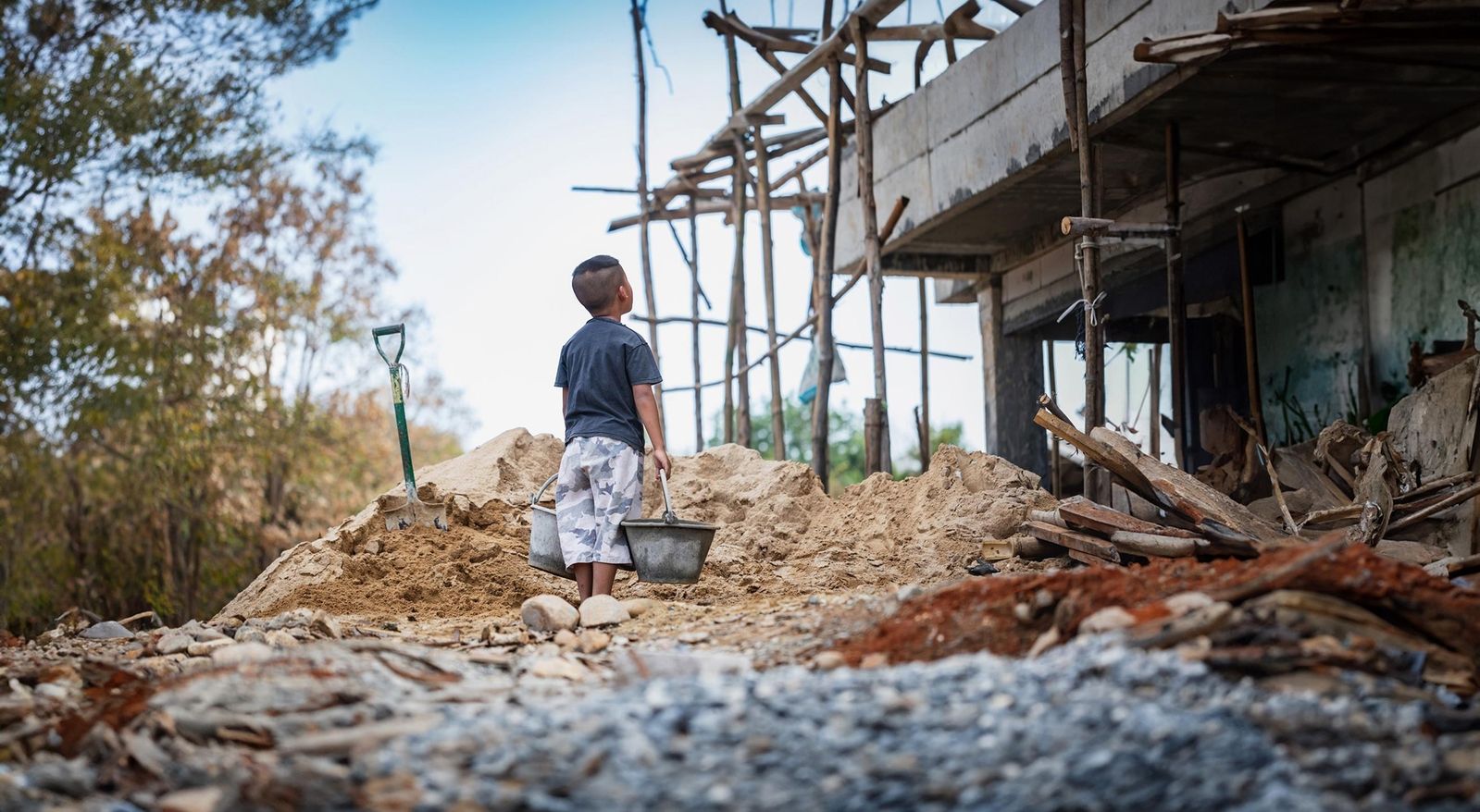 Ein kleiner Junge steht auf einer Baustelle, hält einen Eimer in der Hand und blickt auf ein teilweise fertiggestelltes Gebäude, das von Schutt und Sand umgeben ist.