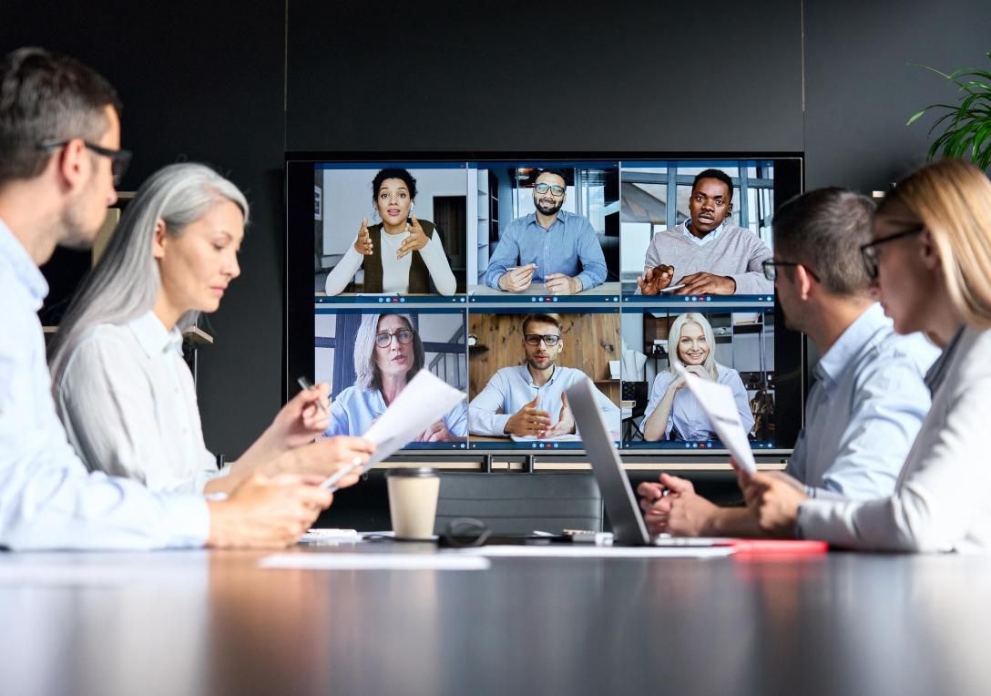 Eine Gruppe von Fachleuten sitzt an einem Konferenztisch und führt einen Videoanruf, der auf einem Bildschirm mit mehreren Remote-Teilnehmern angezeigt wird.